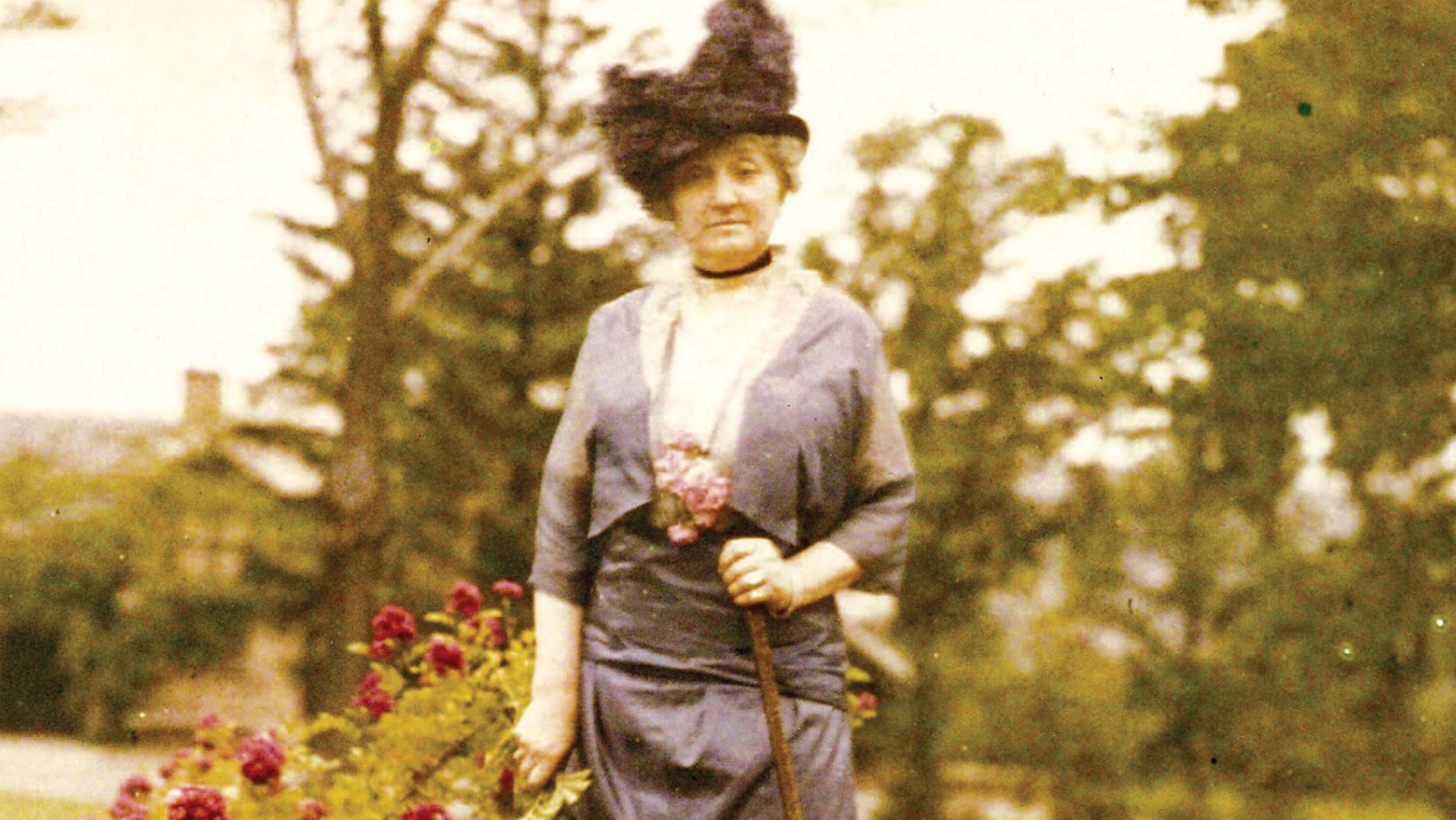 A vintage color photograph of a woman in a gray turn-of-the-century dress and large black hat standing next to red roses.